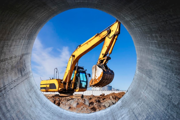 A powerful caterpillar excavator digs the ground against the blue sky Earthworks with heavy equipment at the construction site View through a large concrete pipe