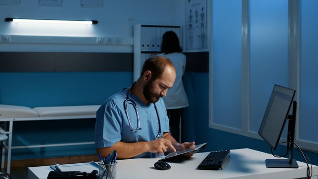 Practitioner assistant sitting at desk holding tablet computer analyzing patient disease report while working late at night in hospital office. Man nurse typing medical expertise. Medicine service