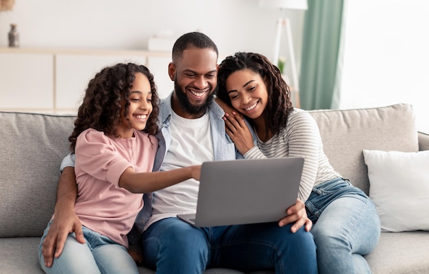 Precious Time With Family. Portrait of happy afro parents and cute little girl using laptop together, watching photos or movie, browsing internet, choosing gifts online sitting on sofa in living room