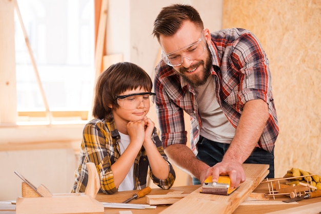 Precision and perfection.Smiling young male carpenter showing his son how to sand wood in his workshop