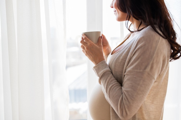 pregnancy, drinks, rest, people and expectation concept - close up of happy pregnant woman with cup drinking tea looking through window at home