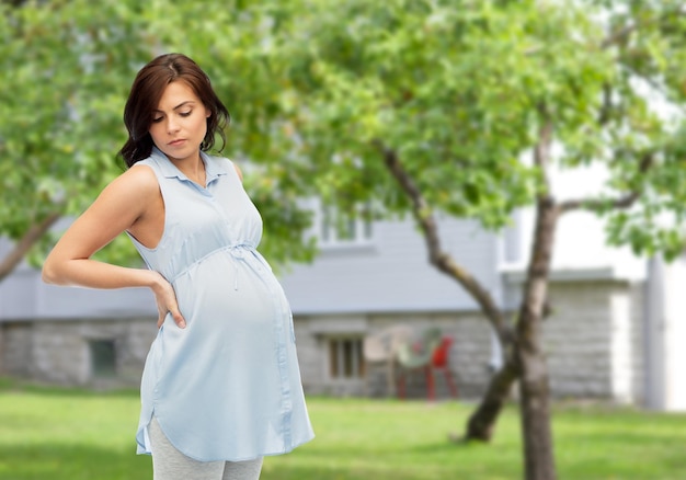 pregnancy, health, people and expectation concept - pregnant woman touching her back and suffering from backache over summer garden and house background