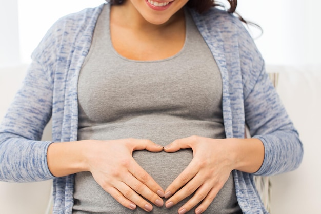 pregnancy, love, care, people and expectation concept - close up of happy pregnant woman making heart gesture at home