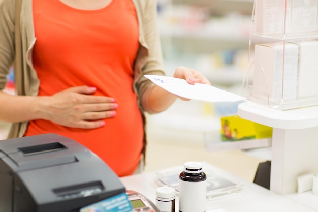 pregnancy, medicine, pharmaceutics, health care and people concept - close up of pregnant woman with prescription buying medication at pharmacy cash register