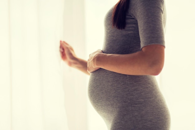 pregnancy, motherhood, people and expectation concept - close up of pregnant woman looking through window at home