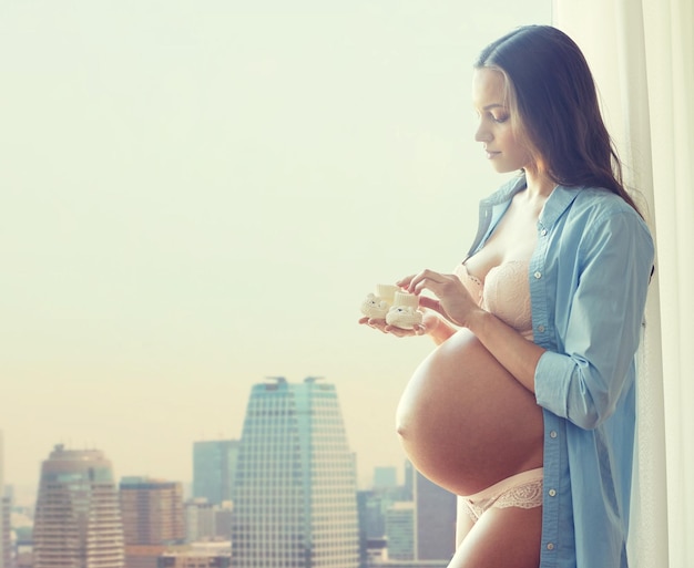 pregnancy, motherhood, people and expectation concept - happy pregnant woman with big bare tummy holding little baby booties at home over city background