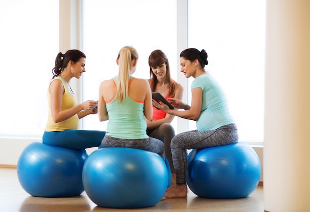 pregnancy, sport, fitness, people and healthy lifestyle concept - group of happy pregnant women with tablet pc computer and smartphone sitting on balls in gym