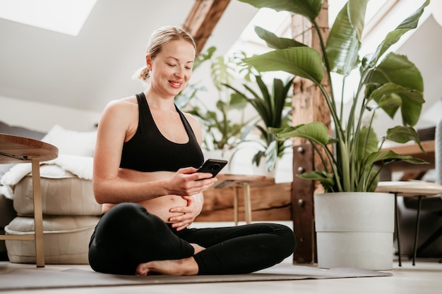 Pregnancy and technoloy supported healthy lifestyle concept Cheerful happy pregnant woman using smart phone application while exercising on yoga mat on living room floor at home