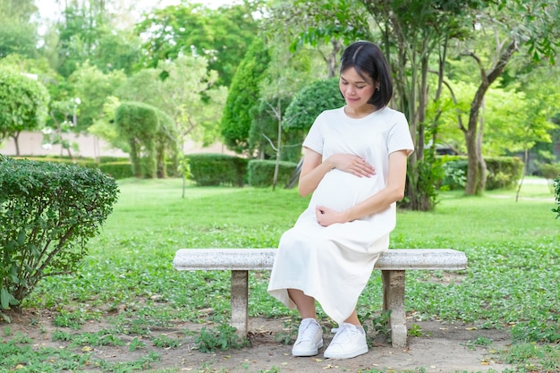 A pregnant Asian woman wearing casual clothes is sitting and looking at her belly at the park.