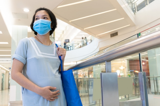 Pregnant Asian woman wearing face mask and touching belly in the shopping mall. New normal life concept.