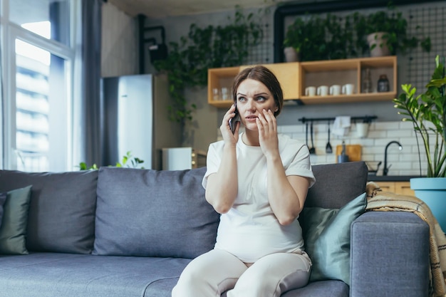 Pregnant beautiful woman sitting on the couch at home talking on a cell phone with a doctor calling an ambulance giving birth