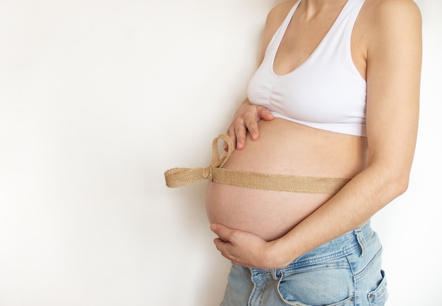 Pregnant belly with eco canvas linen ribbon around the belly on a white wall. Profile view. Place for text