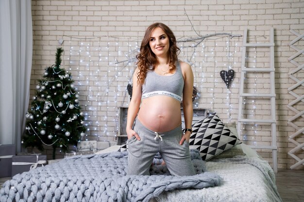 A pregnant brunette in lingerie is sitting on a bed looking at her stomach, happy motherhood. Big gray bed and gray soft armchairs. Christmas tree with toys, New Year's atmosphere