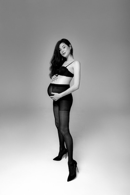 Pregnant brunette woman in pantyhose bra and shoes stands on a gray background