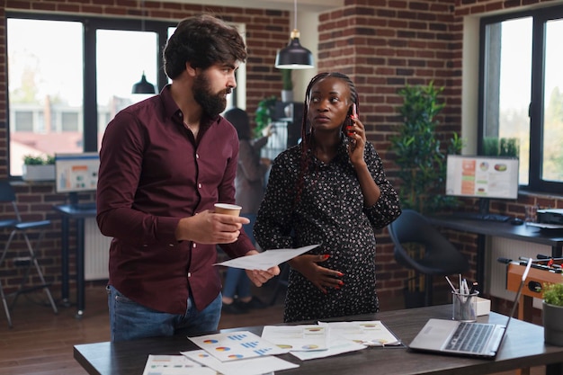 Pregnant businesswoman talking on smartphone while coworker looking over project charts. Multi ethnic diverse people at company job teamworking to solve marketing business issues.