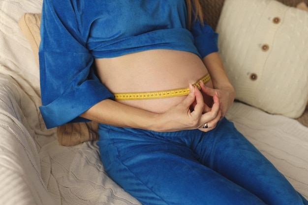 A pregnant girl in a bright blue suit measures her belly with a centimeter tape