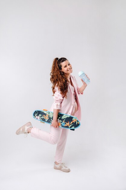 A pregnant girl in a pink suit with a skateboard in her hands drinks juice on a gray background