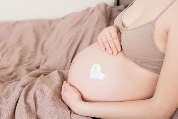 A pregnant girl sits at home on the bed and smears heart an antistretch mark cream on her stomach Pregnancy motherhood preparation and expectation concept