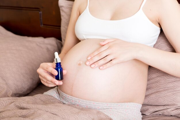 a pregnant girl sits at home on the bed and smears smile an antistretch mark cream on her stomach Pregnancy motherhood preparation and expectation concept