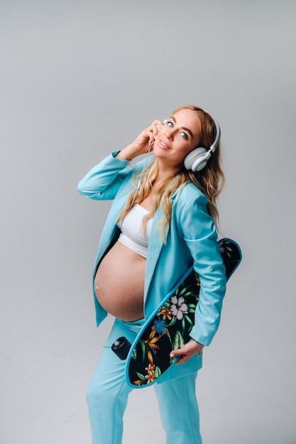 A pregnant girl in a turquoise suit with a skateboard in her hands and headphones stands on a gray background