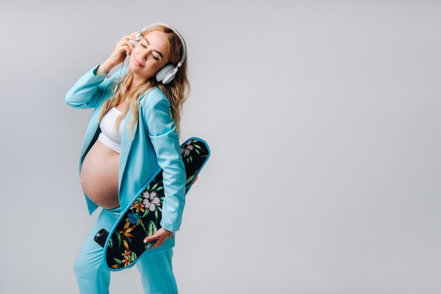 A pregnant girl in a turquoise suit with a skateboard in her hands and headphones stands on a gray background.