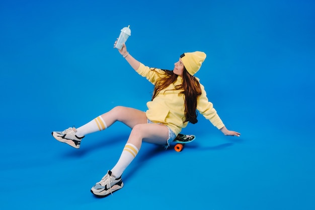 A pregnant girl in yellow clothes with a glass of juice sits on a skateboard on a blue background