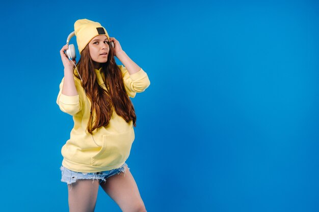 A pregnant girl in a yellow jacket and headphones stands on a blue background