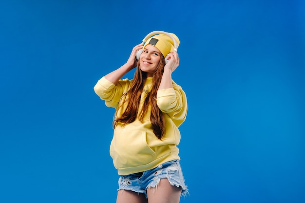 A pregnant girl in a yellow jacket and headphones stands on a blue background