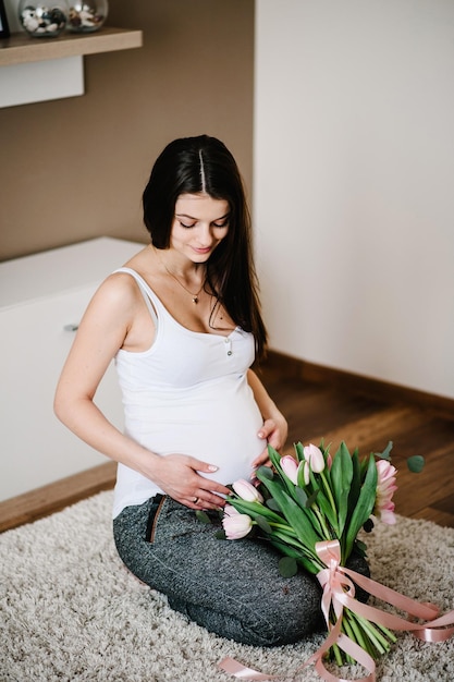 Pregnant happy woman sitting on floor and touching her belly holding a bouquet of flowers hugging tummy at home Baby Shower Motherhood concept Pregnancy rest people expectation