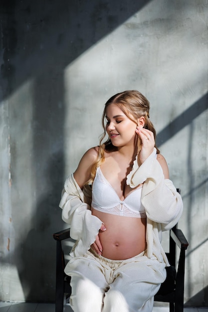 A pregnant pretty woman in a white suit and bra is sitting on a gray background Pregnant cute woman stroking her belly and smiling Happy period of pregnancy