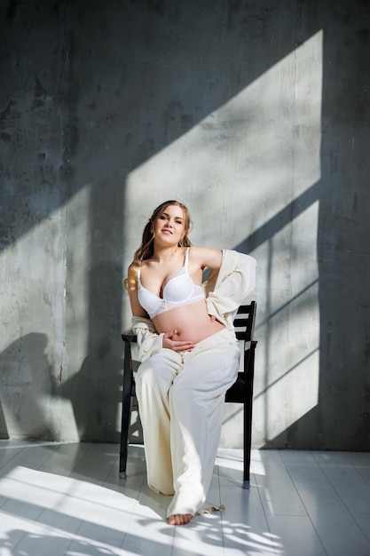 A pregnant pretty woman in a white suit and bra is sitting on a gray background Pregnant cute woman stroking her belly and smiling Happy period of pregnancy