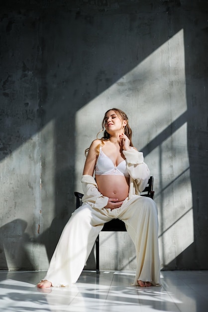 A pregnant pretty woman in a white suit and bra is sitting on a gray background Pregnant cute woman stroking her belly and smiling Happy period of pregnancy