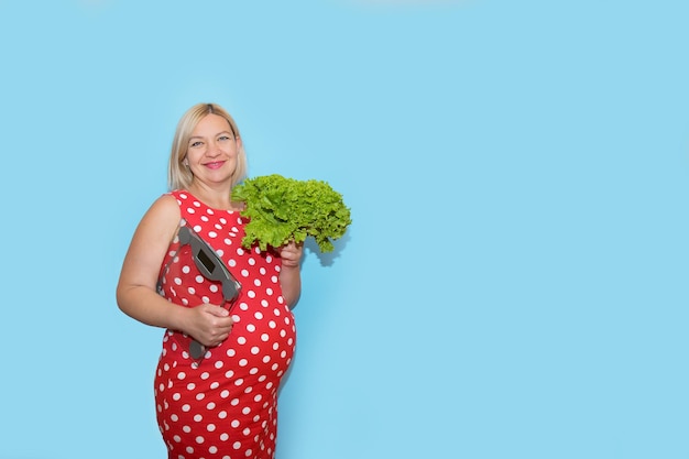 Pregnant smiling woman in a red polka dot dress with a scale in one hand and a salad in the other on a blue background with copy space