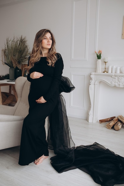 Pregnant woman in black dress sitting on the chair in the studio with white background Pregnant woman touching belly