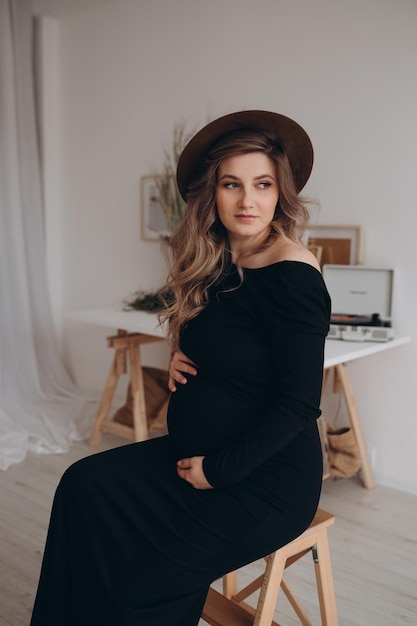 Pregnant woman in black dress sitting on the chair in the studio with white background Pregnant woman touching belly