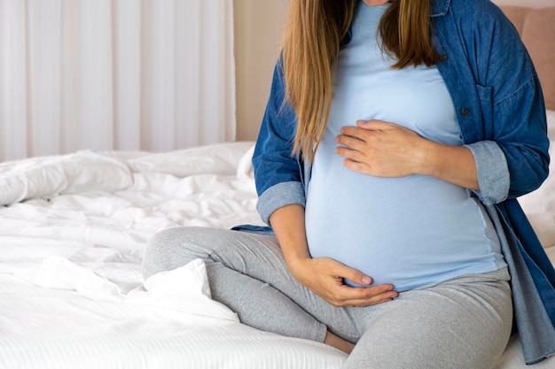 Pregnant woman in blue tshirt and jeans shirt sitting in bed touching her belly Pregnancy maternity preparing and expectation concept Love baby care and parenting