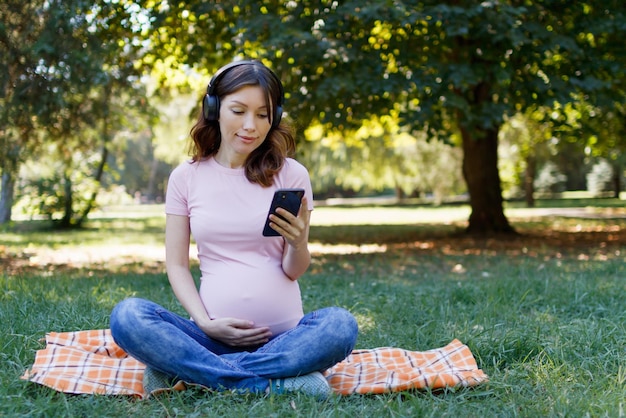 A pregnant woman in headphones listens to music and uses the phone in the park on a summer day