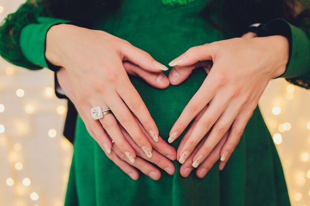 Pregnant Woman and Her Husband holding her hands in a heart shape on her baby bump.