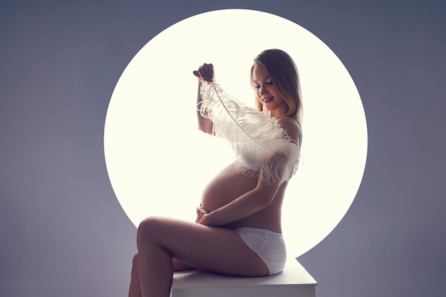 Pregnant woman in her last month of pregnancy in her underwear sits on a cube against white