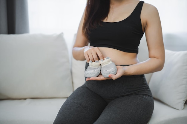 Pregnant woman holding baby shoes on her belly sitting on sofa couch Family mother mom pregnant concept