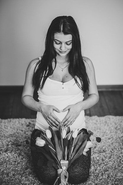 Pregnant woman holding belly with a bouquet of flowers hugging tummy making symbol heart hands at home Baby Shower Motherhood concept Black and white photo