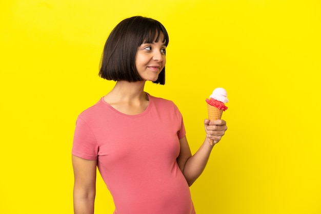 Pregnant woman holding a cornet ice cream isolated on yellow background looking to the side and smiling