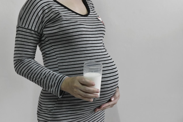 Pregnant woman holding glass milk and hand touching belly on grey background copy space