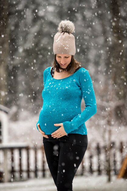 Pregnant woman holding her belly standing outside during snowfall wearing hat and blue sweater