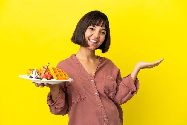 Pregnant woman holding waffles isolated on yellow background extending hands to the side for inviting to come