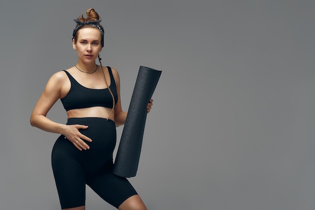 Pregnant woman holding yoga mat in hands going for sport while awaiting baby looking smiling at camera and touching her belly standing against gray wall