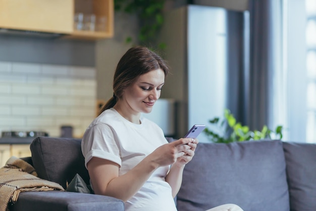 Pregnant woman at home uses the phone while sitting on the couch