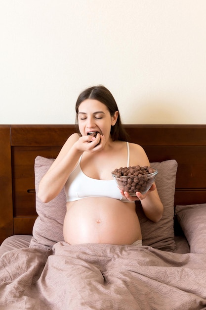 Pregnant woman is eating crunchy chocolate cereal balls from a bowl resting in bed Enjoying food during pregnancy concept