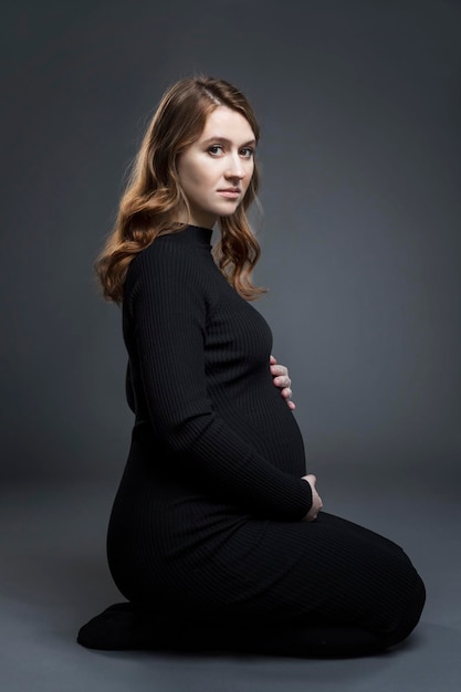 A pregnant woman is sitting on the floor A beautiful young brownhaired woman in a black tightfitting dress Happiness in anticipation of a baby Grey background Profile view Vertical