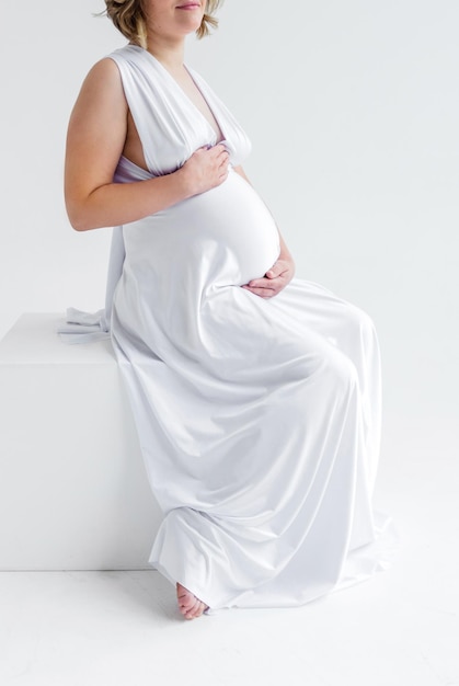 A pregnant woman in a long white silk dress hugs her belly and poses on a white background sitting on a white cube